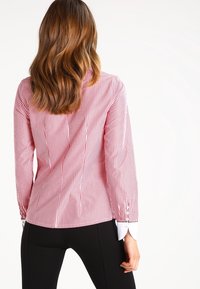 Camisa de botones a rayas rojas y blancas con mangas largas, que presenta un collar blanco y detalles de botones en los puños. Corte entallado.