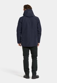Veste imperméable marine avec capuche réglable, manches longues et coupe droite. Portée avec un pantalon vert foncé et des chaussures noires.