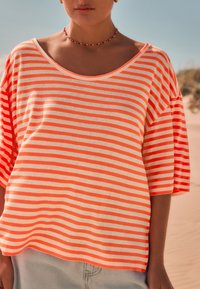 Striped oversized t-shirt in orange and cream cotton, featuring a wide neckline and short sleeves, with a relaxed fit and soft texture.