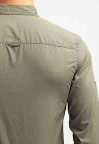 Camicia verde oliva, tessuto leggero, cuciture visibili, dettaglio con bottone sulla spalla, piega sul retro per una maggiore libertà di movimento e texture liscia.