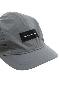 Gorra gris hecha de un tejido ligero con un patrón texturizado. Presenta un parche rectangular negro con texto blanco en el frente.