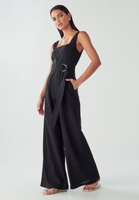 Žena stojící v černém bezramenném širokém overalovém kostýmu s opaskem, doplněném o zlaté náušnice a béžové sandály na podpatku.