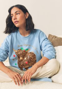 Niebieska bluza z kapturem z nadrukiem misia w stylu kreskówkowym z ozdobami świątecznymi i napisem, materiał o miękkiej fakturze i luźnym kroju.