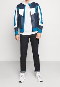 Chaqueta bomber de cuero en azul marino y turquesa con acentos blancos, puños acanalados texturizados y cremallera frontal. Llevada sobre una sudadera gris, combinada con jeans oscuros y zapatillas blancas.
