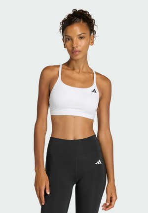 Mujer con cabello rizado que lleva un sujetador deportivo blanco y leggings negros de cintura alta con logos de Adidas, de pie frente a un fondo liso.