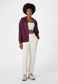 Bordeauxfarbener Kapuzenpullover mit Reißverschluss, weißes bauchfreies Oberteil und cremefarbene Jogginghosen mit elastischen Bündchen. Beige Turnschuhe vervollständigen das Outfit.