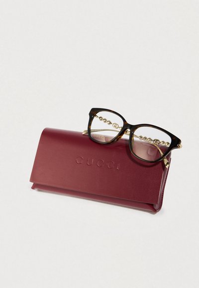 Óculos tartaruga com hastes douradas em contas descansando sobre uma caixa de óculos Gucci bordeaux com logótipo em relevo.