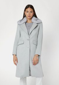 Cappotto in lana grigio chiaro con un morbido colletto in pelliccia sintetica grigia, due tasche frontali e chiusura con un solo bottone. Maniche lunghe e design fino al ginocchio.