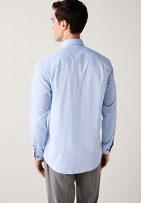 Ljusblå skjorta med lång ärm, mönstrad struktur, knappmanschetter och klassisk krage. Modellen bär grå ränder byxor.