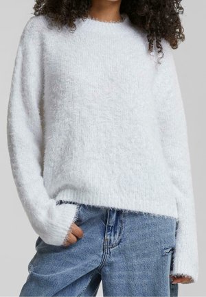 Modèle portant un pull en maille blanc et duveteux à manches longues et un jean bleu, sur un fond gris uni.
