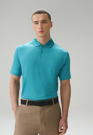 Fiatal férfi rövid hajjal, türkizkék galléros pólót, bézs nadrágot és fekete övet viselve, világosszürke háttér előtt pózol.