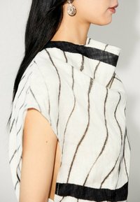 Vêtement en tissu blanc avec des lignes ondulées noires, doté d'un col haut et de manches courtes. Des accents texturés et des côtés ouverts viennent enrichir le design.