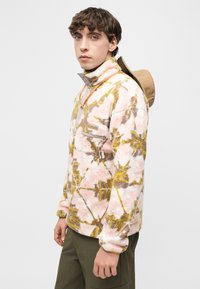 Νέος άνδρας που φοράει ροζ και κίτρινο φλις μπουφάν με ελιά παντελόνι και μπεζ καπέλο κρεμασμένο στην πλάτη του, στέκεται μπροστά σε λευκό φόντο.