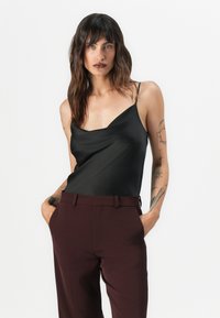 Schwarzes Satin-Camisole mit dünnen Trägern, drapierter Schnitt, getragen mit hochgeschnittenen burgunderfarbenen Hosen. Glatte Textur, minimalistischer Stil.