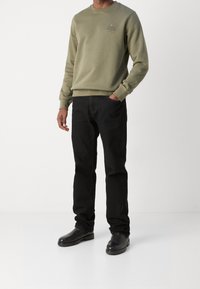Uomo che indossa un sudore oliva di Tommy Hilfiger, jeans neri e stivali di pelle neri, in piedi con una mano in tasca.