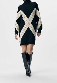 Gebreide jurk in zwart met crèmekleurige diagonale strepen, met een hoge col en losse mouwen. Gepaard met zwarte leren laarzen tot de knie.