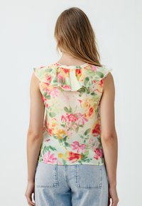 Blumiges ärmelloses Oberteil mit Rüschenkragen, in sanften Farben, aus weichem Stoff und mit einem leicht luftigen Design. Kombiniert mit blauen Jeans.