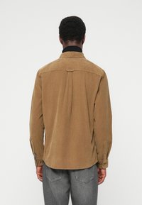 Chemise en velours côtelé marron avec col et poignets boutonnés, présentant une texture côtelée verticale et un pli central à l'arrière.