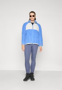 Blå fleecejakke med en kremfarget panel, glidelås foran og høy krage, kombinert med grå leggings og svarte støvler. Solbriller fullfører stilen.