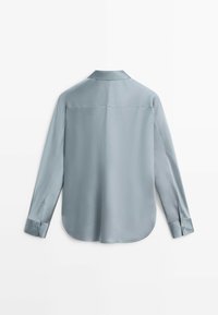 Camicia azzurra a maniche lunghe con colletto, polsini con bottoni e orlo curvo vista da dietro su sfondo bianco.