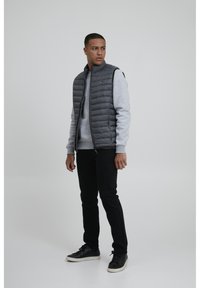 Chaleco acolchado gris sobre un sudadera gris claro, jeans negros y zapatillas negras. El chaleco tiene un cierre frontal y detalle de costuras horizontales.