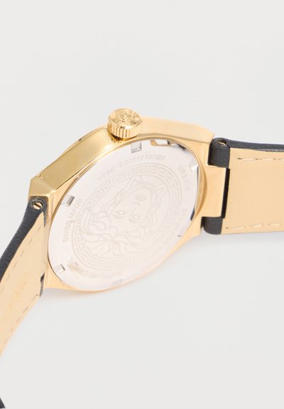 Πίσω πλευρά χρυσού ρολογιού με χαραγμένο σχέδιο κεφαλής Μέδουσας, μαύρο δερμάτινο λουράκι με μπεζ εσωτερικό και χρυσό κορώνα με λογότυπο.