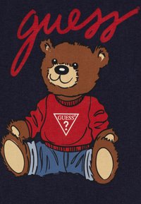 Gráfico de un oso pardo con un suéter rojo con un logo de triángulo blanco, jeans azules, contra un fondo marino con "guess" en letras rojas.