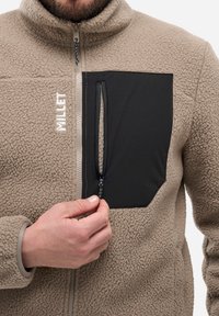 Homme portant une veste en polaire beige avec une poche poitrine zippée noire, tirant sur le tirette de la fermeture éclair, logo "MILLET" brodé sur le devant.