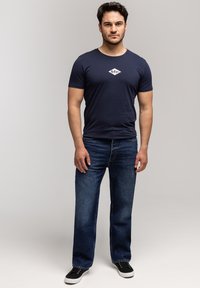 Mężczyzna stoi w granatowej koszulce z białym logo, ciemnoniebieskich dżinsach i czarnych sneakersach na jednolitym jasnym tle.