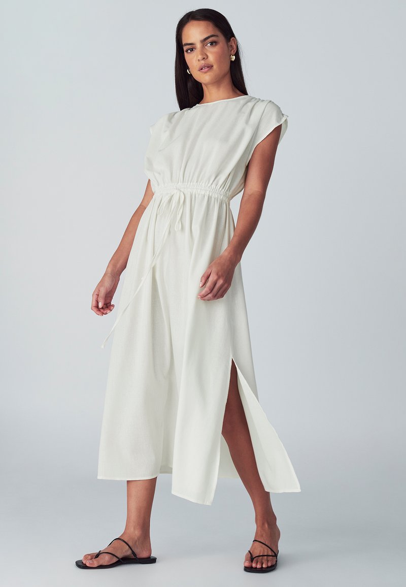 Bílá midi šaty s krátkým rukávem, elastickým pasem, bočními rozparky a hladkou texturou. Nosené s černými plochými sandály.