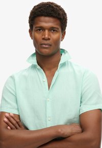 Camicetta verde menta a maniche corte con colletto, caratterizzata da una texture sottile e chiusura con bottoni, indossata da un modello con le braccia incrociate.