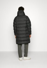 Crna puffer jakna s horizontalno qiltanim dijelovima, koja ima kapuljaču i dugi dizajn. Uparena s sivim hlačama i bijelim tenisicama.