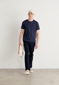 Navy kortermet t-skjorte med brystlomme, kombinert med mørke bukser og en kremfarget genser. Beige sneakers; modellen har på seg en lys caps.