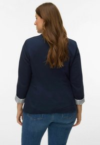 Marineblauwe jas met een getailleerde pasvorm en driekwart mouwen, met een gestreept manchetdetail. Draagt bij blauwe spijkerbroek.