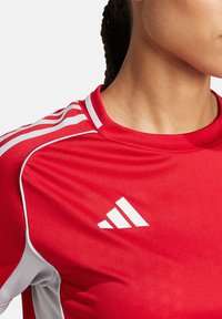 Rode sportshirt met witte accenten en drie verticale strepen op de schouders, met een prominent wit Adidas-logo.