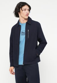 Jūras spēku zip-up jaka ar apkakli, izgatavota no auduma, ar sānu kabatām un ar rāvējslēdzēju slēgtu krūšu kabatu, apģērbs gaismas zilā T-kreklā.