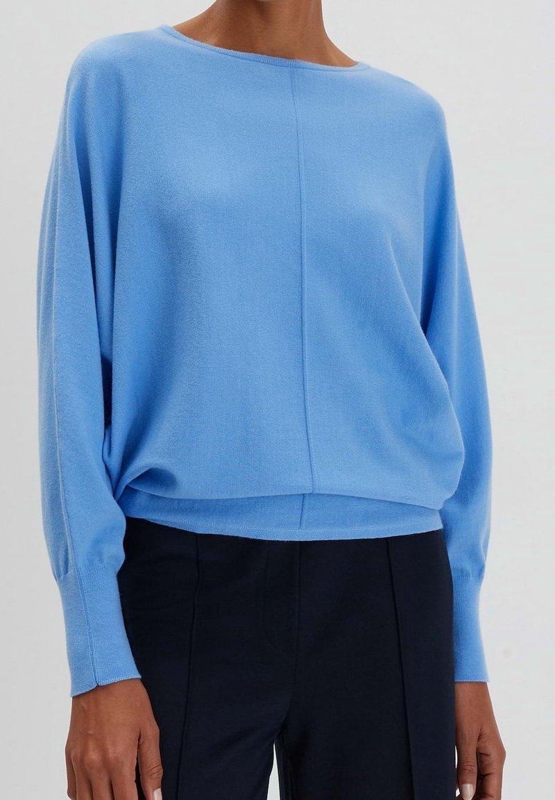 Femme portant un pull bleu clair à manches longues avec une couture centrale, associé à un pantalon noir taille haute sur un fond uni.