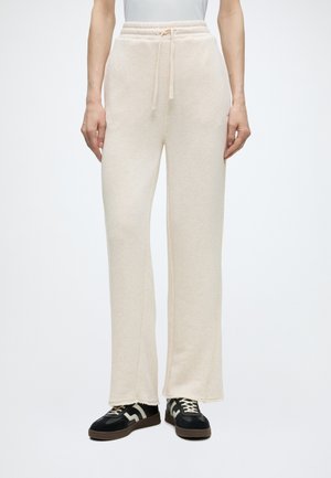 Person iført cremefarvede sweatpants med vide ben og snøre i taljen samt sorte sneakers med hvide detaljer, stående mod en ensfarvet baggrund.