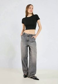 Musta lyhythihainen crop-top, yhdistettynä vaaleanharmaisiin leveälahkeisiin farkkuihin, joissa on haalistunut pinta. Malli seisoo kädet taskuissa.