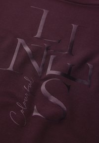 Burgunderfarbener Stoff mit großen, stilisierten schwarzen Buchstaben, die "LINES" schreiben, mit dem Wort "Farblich" in einer kleineren, geschwungenen Schrift darunter.