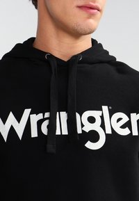 Felpa nera con un grande logo bianco "Wrangler" sul davanti. Presenta un cappuccio con cordino e un tessuto morbido e testurizzato.