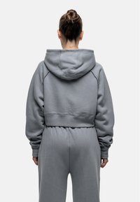 Felpa corta grigia realizzata in tessuto morbido, con cappuccio, vestibilità rilassata e vita a coste. Il design include cuciture sulle spalle scese.