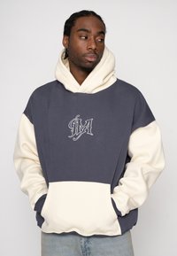 Sweatshirt met capuchon in marineblauw en crèmekleur, voorzien van een voorzak, geborduurd logo en contrasterende mouwen met een zachte textuur.