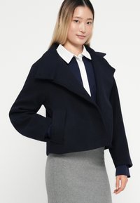 Marineblå uld-blandings kort jakke med stor krave, skrå lommer og en struktureret design. Parret med en hvid skjorte og en grå nederdel.