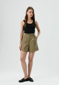 Khaki shorts met hoge taille en voorplooien, opgerolde boorden, gecombineerd met een zwart tanktop en zwarte loafers. Soepele stof en een nauwkeurige pasvorm.
