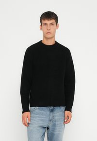 Zwarte geribde gebreide trui met een ronde halslijn en lange mouwen, gecombineerd met lichtblauwe jeans. Gladde textuur, casual fit.