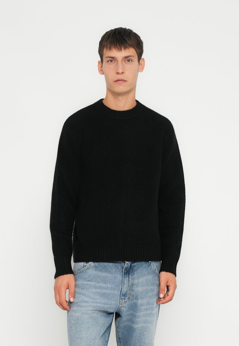 Zwarte geribde gebreide trui met een ronde halslijn en lange mouwen, gecombineerd met lichtblauwe jeans. Gladde textuur, casual fit.