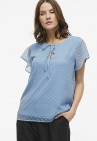 Blusa azzurro chiaro con maniche corte in chiffon trasparente a sbuffo, texture a pois, scollatura a chiave e dettaglio con fiocco annodato.