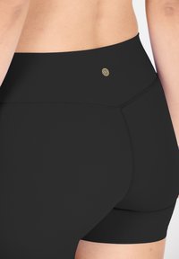 Shorts noirs taille haute en matériau extensible avec une texture lisse. Présente un petit emblème doré et des coutures plates pour le confort.