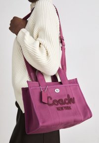 Purple canvas tote bag with embossed "Coach New York" logo, double handles, and a detachable tag, worn on a person in a knit sweater.